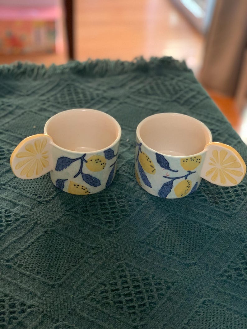 Pode incluir: Duas canecas de cer&acirc;mica com uma al&ccedil;a em forma de fatia de lim&atilde;o. As canecas t&ecirc;m um fundo azul claro com lim&otilde;es amarelos e folhas azuis. O interior das canecas &eacute; branco. As canecas est&atilde;o sobre uma superf&iacute;cie verde texturizada.