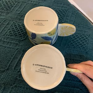 Pode incluir: Duas canecas de cer&acirc;mica com al&ccedil;as em forma de fatia de lim&atilde;o e desenhos de lim&otilde;es e flores. As canecas s&atilde;o brancas com detalhes amarelos e azuis. A parte inferior das canecas diz "by ANTHROPOLOGIE" e "Made in China".