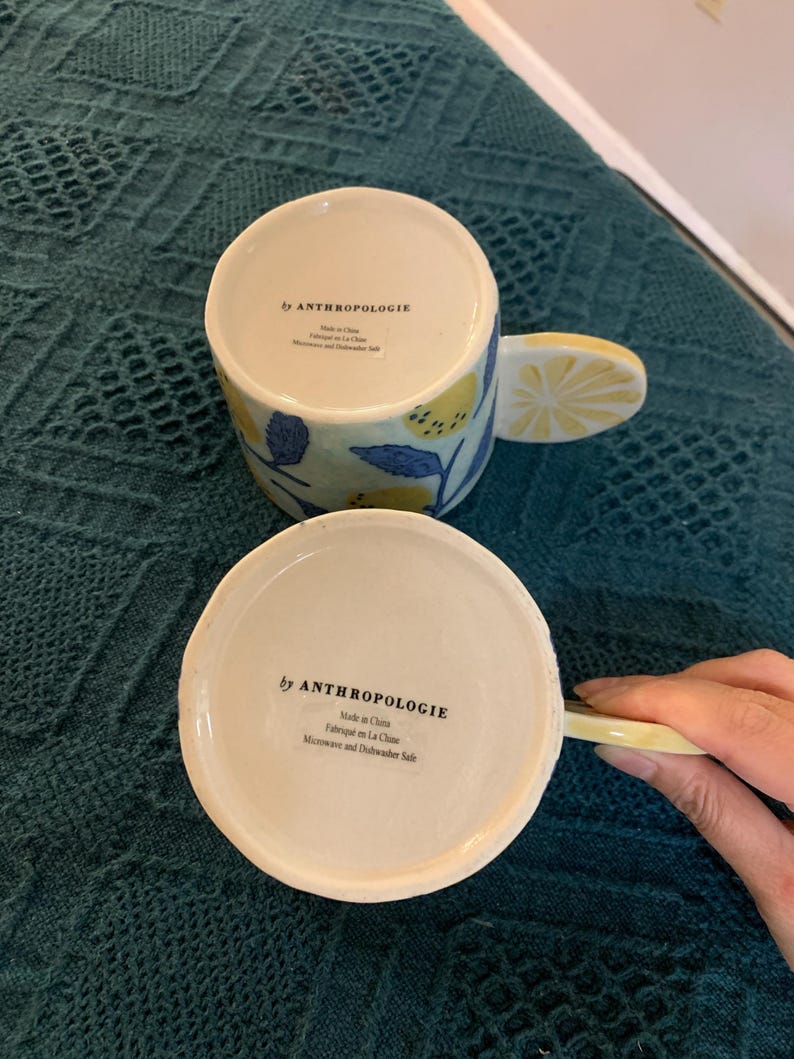 Pode incluir: Duas canecas de cer&acirc;mica com al&ccedil;as em forma de fatia de lim&atilde;o e desenhos de lim&otilde;es e flores. As canecas s&atilde;o brancas com detalhes amarelos e azuis. A parte inferior das canecas diz "by ANTHROPOLOGIE" e "Made in China".