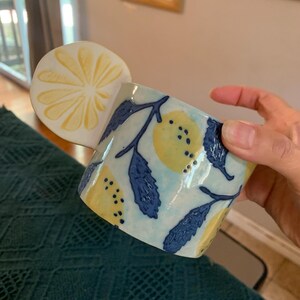 Pode incluir: Uma caneca de cer&acirc;mica com uma pega em forma de fatia de lim&atilde;o. A caneca apresenta um fundo azul claro com lim&otilde;es amarelos e folhas azuis escuras. A caneca &eacute; segurada por uma m&atilde;o.