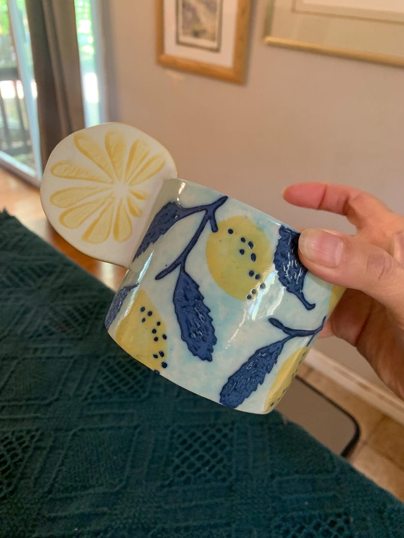 Pode incluir: Uma caneca de cer&acirc;mica com uma pega em forma de fatia de lim&atilde;o. A caneca apresenta um fundo azul claro com lim&otilde;es amarelos e folhas azuis escuras. A caneca &eacute; segurada por uma m&atilde;o.
