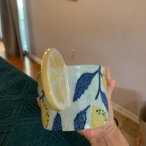 Pode incluir: Uma caneca de cer&acirc;mica com uma al&ccedil;a de fatia de lim&atilde;o e um padr&atilde;o de lim&atilde;o e folha. A caneca &eacute; azul clara com lim&otilde;es amarelos e folhas azuis escuras. A caneca &eacute; segurada por uma pessoa.