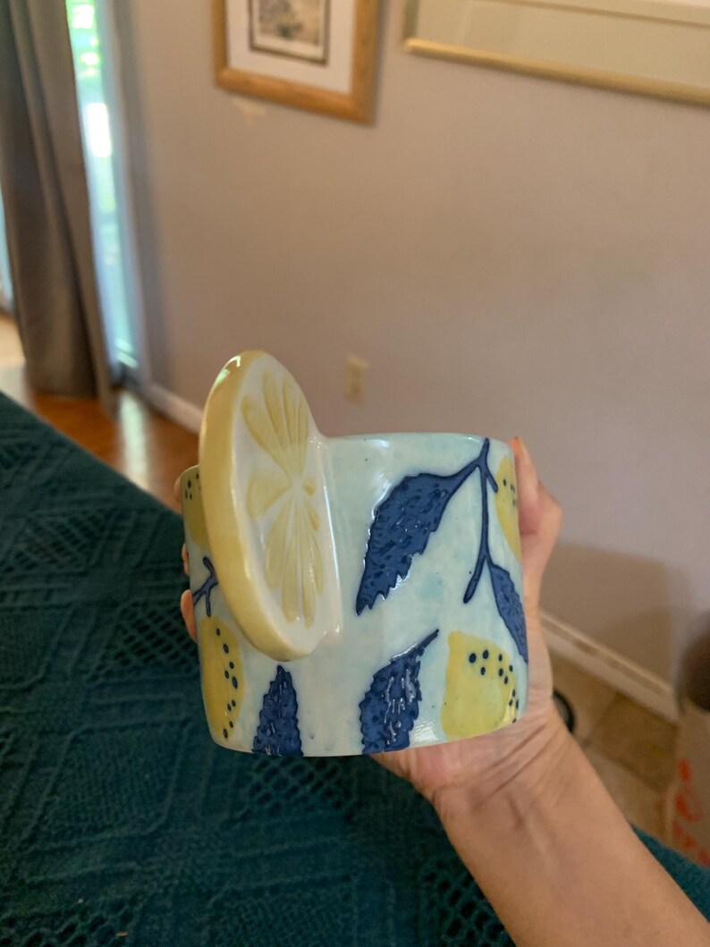 Pode incluir: Uma caneca de cer&acirc;mica com uma al&ccedil;a de fatia de lim&atilde;o e um padr&atilde;o de lim&atilde;o e folha. A caneca &eacute; azul clara com lim&otilde;es amarelos e folhas azuis escuras. A caneca &eacute; segurada por uma pessoa.