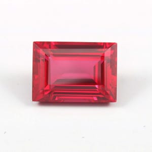 May include: A vibrant, rectangular-cut gemstone with a deep, saturated red hue. The stone features a faceted surface that creates sharp geometric reflections and a clear, transparent appearance, showcasing a classic emerald-style cut design.