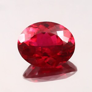 May include: A faceted, oval-shaped ruby gemstone. The gem is a deep, saturated red color with a high degree of clarity. The ruby is set against a white background, with its reflection visible below. The gemstone is a symbol of love and passion.