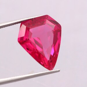 May include: A vibrant, deep pink gemstone with a shield-like, faceted cut. The stone is held by silver-toned metal tweezers, showcasing its translucent quality and sharp, geometric edges. The facets reflect light, creating a brilliant appearance.