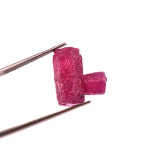 Puede incluir: Dos piedras preciosas de rubí en bruto sin cortar sujetas con pinzas sobre un fondo blanco. Los rubíes son de un rojo intenso y vibrante con una superficie texturizada. Uno es más grande y alargado que el otro.