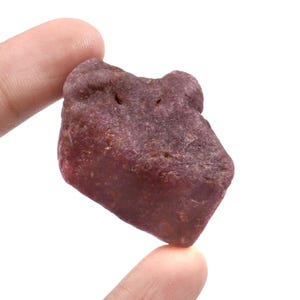 Pode incluir: Uma gema de rubi bruta e não cortada, segurada entre dois dedos. O rubi é de uma cor vermelha profunda e rica com uma superfície texturizada. A gema tem uma forma irregular.