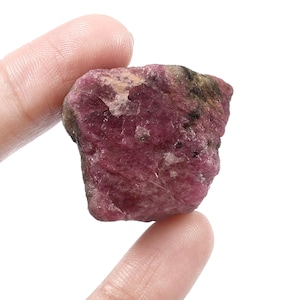 Puede incluir: Una gema de rubí en bruto, sin cortar, con un tono rojo-rosado intenso. La piedra natural tiene una superficie texturizada con áreas más claras y oscuras, sostenida entre dos dedos sobre un fondo blanco.