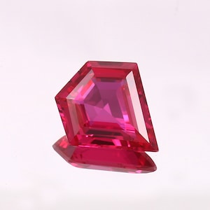 Könnte beinhalten: Ein facettierter, Drachen-förmiger Edelstein in einem leuchtenden Rotton. Der Edelstein hat ein symmetrisches Design mit scharfen Winkeln und eine reflektierende Oberfläche, die ein Spiel von Licht und Schatten erzeugt. Der Edelstein wird auf einer weißen Oberfläche präsentiert.