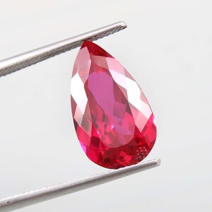 May include: A pear-shaped, faceted gemstone with a vibrant, deep red colour. The stone exhibits high clarity and brilliance, with multiple triangular and kite-shaped facets reflecting light across its surface, held by silver-toned metal tweezers.