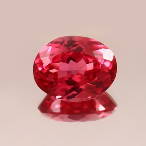 Puede incluir: Una gema facetada, de forma ovalada, con un tono rojo intenso y vibrante. La gema es muy reflectante, con múltiples facetas que capturan la luz. La piedra se encuentra sobre un fondo claro, con su reflejo visible debajo.