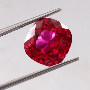 May include: A vibrant, cushion-cut red gemstone held by silver-toned metal tweezers. The stone features a faceted surface that reflects light, creating a deep, saturated crimson color with bright highlights across its polished, geometric facets.
