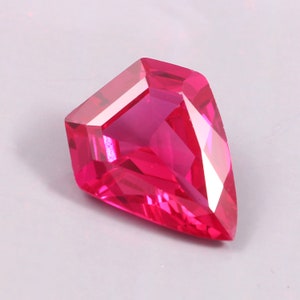 May include: A vivid pink, kite-shaped gemstone with multiple facets. The gem's cut creates a sparkling effect as it reflects light. The gemstone is set against a plain, light background, highlighting its colour and shape.