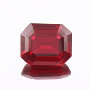 May include: A vibrant, deep red gemstone cut into a rectangular emerald shape with sharp, clean facets. The stone displays high clarity and a rich, saturated crimson hue, reflecting light across its polished surface with a clear mirror reflection.