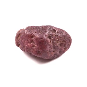 Puede incluir: Una gema de rubí natural, de color rojo púrpura intenso. La piedra tiene una forma irregular y redondeada con una superficie texturizada, sobre un fondo blanco.