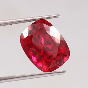May include: A faceted, cushion-cut ruby gemstone held by metal tweezers. The ruby is a deep, vibrant red color with multiple facets reflecting light. The gemstone is a square shape with rounded corners.