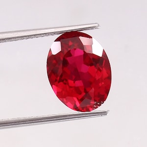 Könnte beinhalten: Ein facettierter, oval geformter Rubin-Edelstein, der von einer Pinzette gehalten wird. Der Rubin hat eine tiefe, leuchtend rote Farbe mit mehreren Facetten, die das Licht reflektieren. Der Edelstein ist ein Blickfang vor einem schlichten weißen Hintergrund.