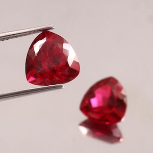 Pode incluir: Duas pedras preciosas vermelhas escuras, facetadas em forma triangular. Uma é segurada por pinças de prata, a outra repousa sobre uma superfície reflexiva. As pedras têm uma aparência brilhante e polida, mostrando a refração interna da luz.