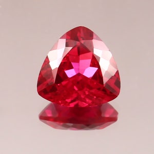 May include: A triangular-cut gemstone in a vivid red colour. The facets catch the light, producing a sparkling effect. The gem is presented on a reflective surface, accentuating its brilliance. The image showcases the gem's clarity and rich colour, a striking display.