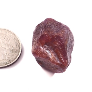 Pode incluir: Uma gema de rubi bruta, não cortada, de cor vermelha escura. O rubi está ao lado de uma moeda de um quarto de dólar dos EUA para comparação de tamanho. A gema tem uma forma natural irregular e uma superfície ligeiramente brilhante. É um mineral natural.