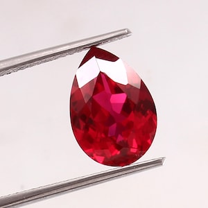 Könnte beinhalten: Ein facettierter, birnenförmiger Rubin-Edelstein, der von einer silbernen Pinzette gehalten wird. Der Rubin hat eine tiefe, leuchtende rote Farbe, mit mehreren Facetten, die das Licht einfangen. Der Edelstein wird vor einem schlichten, weißen Hintergrund präsentiert.