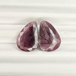 May include: Two pear-shaped pink gemstones with a faceted cut. The gemstones are set against a white background.