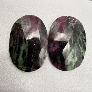 May include: Two oval-shaped green, black and pink gemstones with a faceted cut.