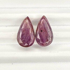 May include: Two pear-shaped gemstones, both a deep reddish-purple colour. The gemstones are faceted and have a slight sheen.