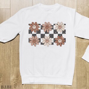 May include: White sweatshirt with a checkered pattern and brown flowers with smiley faces.