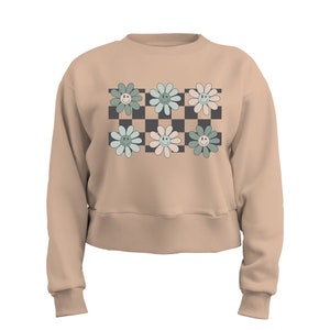 May include: A beige cropped sweatshirt with a checkered pattern in black and white. Six pastel green, blue, and pink flowers with smiley faces are arranged on the checkered pattern.