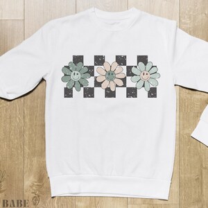 May include: White long-sleeved sweatshirt with a checkered pattern and three smiling flower designs in green, pink, and blue.