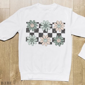 May include: White sweatshirt with a checkered pattern and green, pink, and blue flowers with smiley faces.