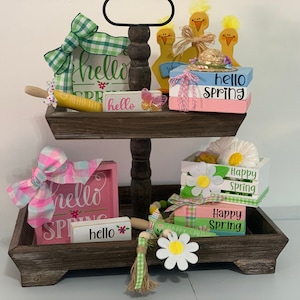 May include: A two-tiered wooden tray decorated with spring-themed items. The top tier features a green and white gingham bow, a rolling pin, and signs that say "hello" and "hello SPRING". The bottom tier has a pink and blue bow, a daisy, and signs that say "Happy Spring".