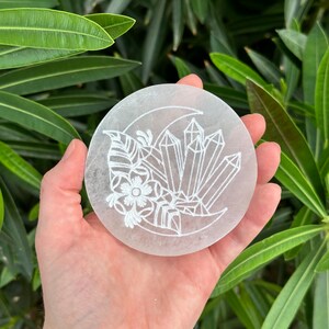 May include: White selenite charging plate with a crescent moon and crystal design. The plate is etched with a floral design and a crescent moon with crystals.