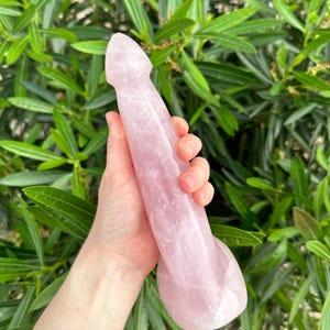 May include: A pink crystal wand, held against a backdrop of green foliage. The wand is a light rose color and has a smooth, polished surface. The shape is elongated with a rounded tip and a flared base.