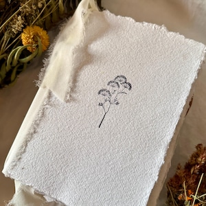 Puede incluir: Un diario rectangular con una cubierta blanca texturizada que presenta un dibujo lineal negro de una flor silvestre. El libro tiene bordes irregulares y está encuadernado con una cinta de tela color crema atada en un nudo en la parte superior.