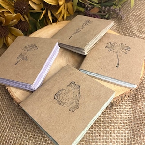 Op de afbeelding: Vier kleine, vierkante notitieboekjes met kraftpapier covers, elk met een andere zwarte botanische illustratie: een bloem, een takje, een madeliefje en een pad. De notitieboekjes zijn gestapeld op een houten schijf, met gedroogde bloemen op de achtergrond.