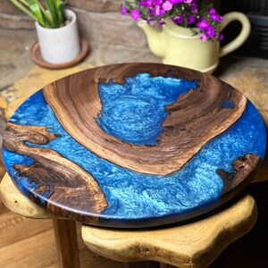 May include: A round wooden serving tray with a blue epoxy resin inlay. The tray has a natural wood finish and a unique, organic shape.