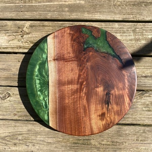 May include: A round wooden serving board with a green epoxy resin inlay. The board has a natural wood finish and the resin has a marbled effect.