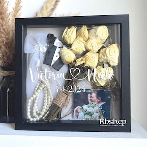 May include: A black framed shadow box with a dried flower bouquet, a white dress, a black tuxedo, a string of pearls, a photo of a couple, and the text "Victoria & Nick Est 2024".