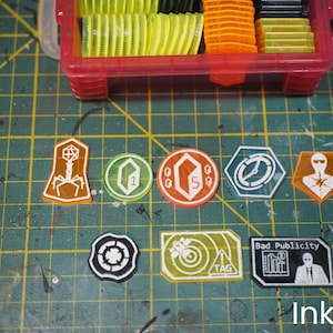 May include: A selection of colourful game tokens and a red plastic container filled with more tokens. The tokens are various shapes, including circles, hexagons, and rectangles, with white and orange graphics. The text "Bad Publicity" is visible on one token.