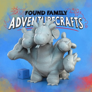 May include: A gray, multi-headed dragon plush toy with a blue die. The text "FOUND FAMILY ADVENTURE CRAFTS" is above the dragon. The dragon has multiple heads, each with a unique horn or ear design.