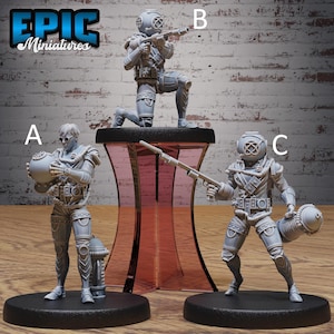 May include: Three grey miniature figures of divers in vintage diving suits. One figure kneels with a rifle, another holds a large sphere, and the third stands with a rifle and a cylindrical object. The figures are on black bases.