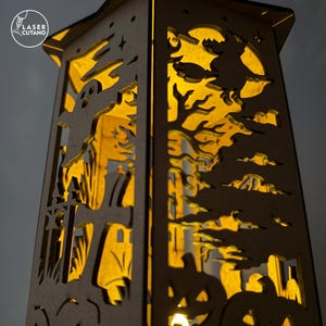 May include: A wooden Halloween lantern with intricate cut-out designs. The lantern features a spooky scene with a ghost, witch, pumpkins, and a full moon. The interior glows with a warm, inviting light, creating a festive ambiance. The text "LASER CUTANO" is in the upper left corner.