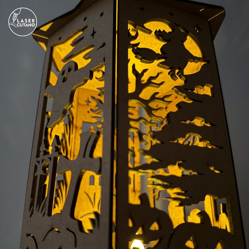 May include: A wooden Halloween lantern with intricate cut-out designs. The lantern features a spooky scene with a ghost, witch, pumpkins, and a full moon. The interior glows with a warm, inviting light, creating a festive ambiance. The text "LASER CUTANO" is in the upper left corner.