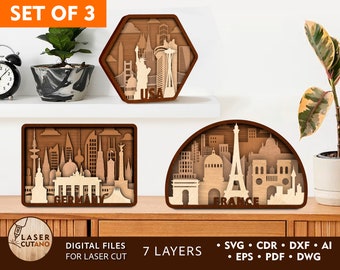May include: Set of three laser cut wooden city skyline designs. The designs are of Berlin, Paris, and New York City. Each design has multiple layers and is labeled with the city name. The designs are on a wooden shelf with a plant and a clock in the background.