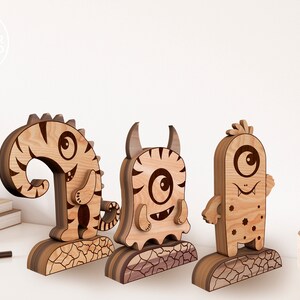Może przedstawiać: Trzy drewniane figurki potwor&oacute;w w paski z jednym dużym okiem. Każdy potw&oacute;r ma inny wyraz twarzy i unikalny design. Potwory stoją na drewnianej podstawie.