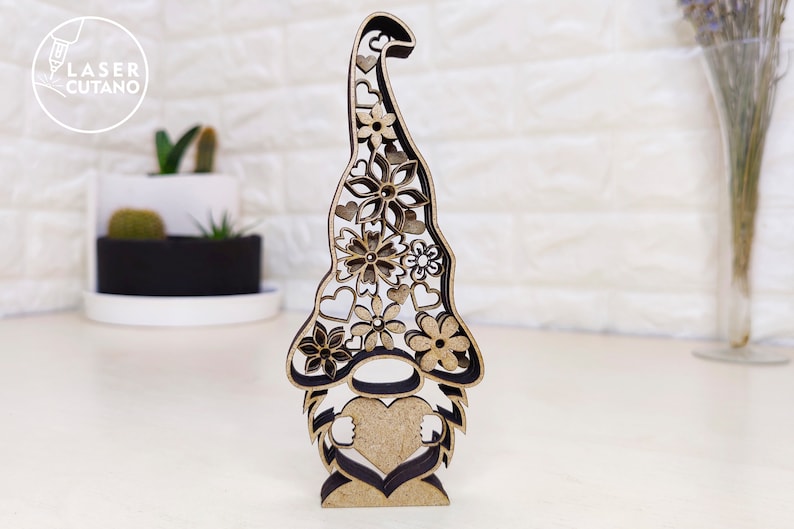 Pu&ograve; includere: Una statuetta di gnomo in legno con un corpo a forma di cuore e un cappello decorato con fiori e cuori. Lo gnomo &egrave; in piedi su una superficie bianca.