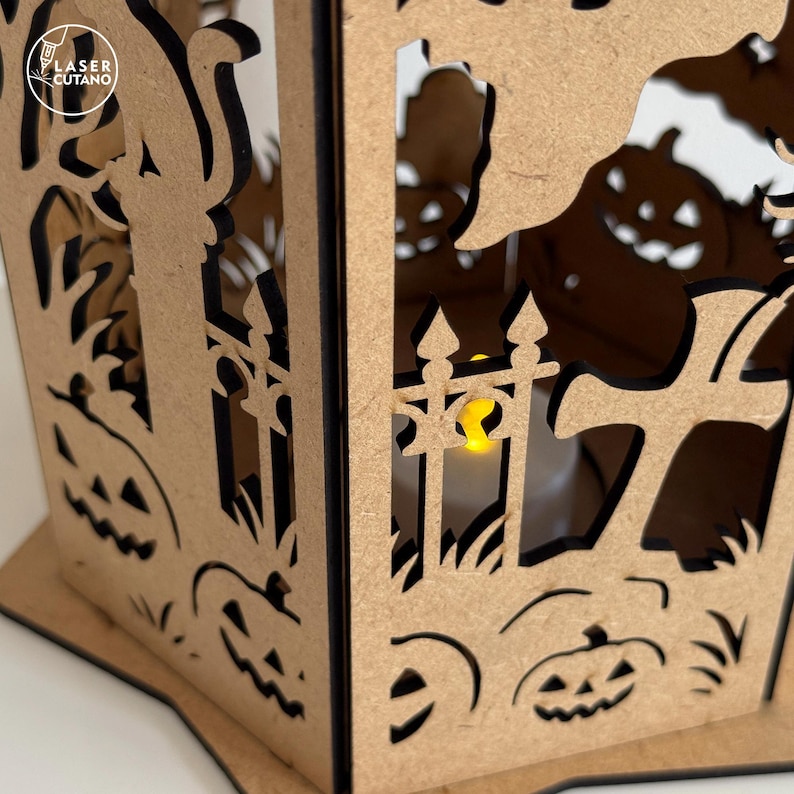 May include: A laser-cut wooden Halloween lantern with a spooky design. The lantern features cutouts of pumpkins, a graveyard scene, and a fence. The lantern has a warm, yellow light inside, creating a festive glow. The text "LASER CUTANO" is visible.